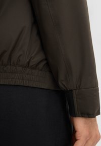 Dunkelgrüne Jacke mit elastischem Saum und Bündchen, aus einem glatten, strukturierten Stoff. Schwarzes Unterhemd ist unter der Jacke sichtbar.