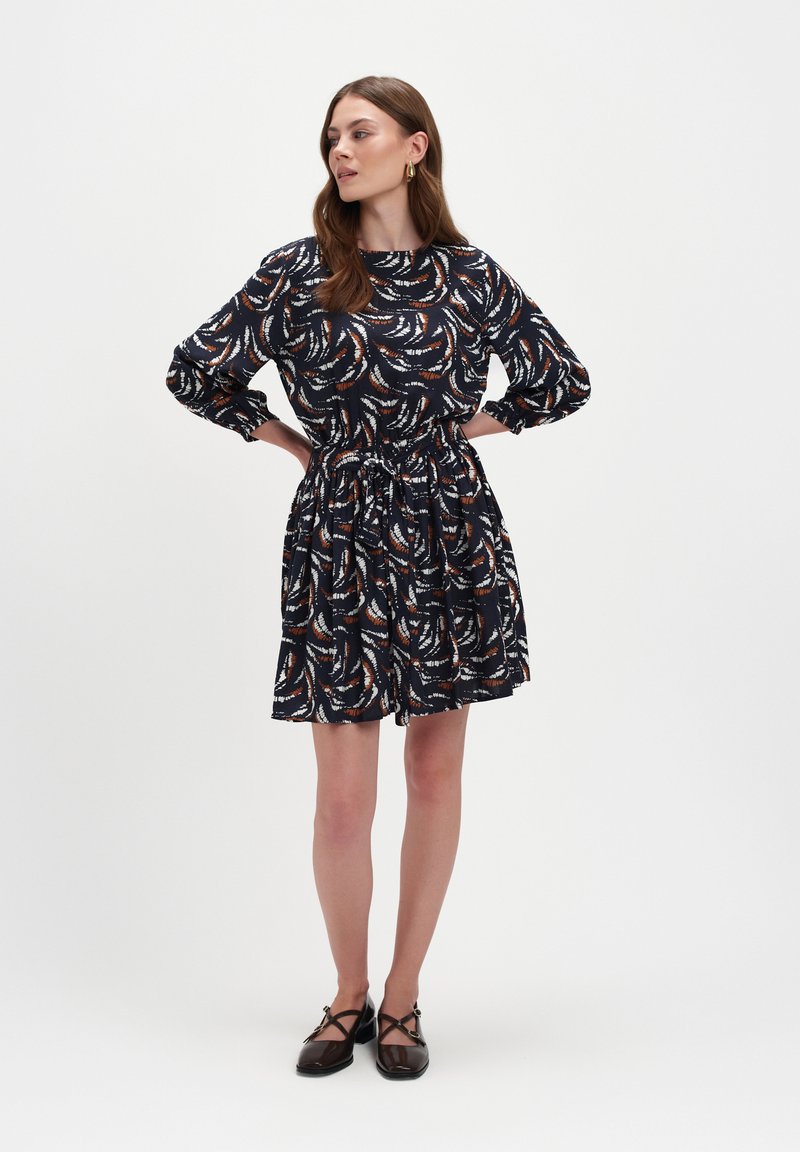 Kobieta stoi z rękami na biodrach, ubrana w czarną sukienkę w brązowe i białe abstrakcyjne wzory oraz czarne buty na krzyż na białym tle.