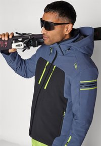 Chaqueta de esquí en azul oscuro y negro con acentos en verde lima. Presenta un cierre de cremallera, capucha y detalles a rayas en las mangas. Textura suave.
