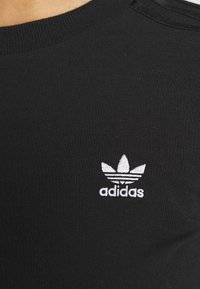 Černé bavlněné tričko s bílým vyšitým logem Adidas, které obsahuje tři pruhy a trojlístkový design na levé části hrudi.