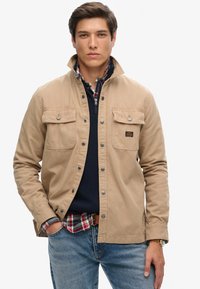 Khaki jack met drukknopen, twee borstzakken en een kraag, gelaagd over een marineblauwe trui en een geruit shirt, gedragen met blauwe jeans.