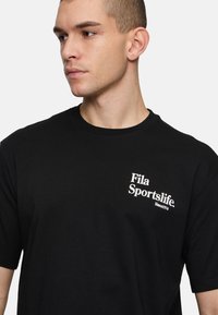 Camiseta de algodón negra con cuello redondo y mangas cortas, con un logo blanco de "Fila Sportslife" en el área del pecho izquierdo.