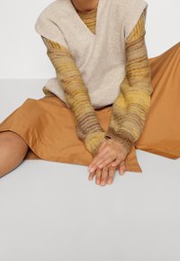 Haut sans manches beige texturé superposé à une chemise multicolore à manches longues. Le mannequin porte une jupe marron et est assis sur une surface grise.