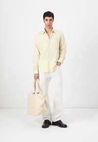 Cămașă galben deschis cu mânecă lungă, dintr-un material texturat, cu un buzunar frontal, asortată cu pantaloni albi și o geantă de umăr bej. Pantofii negri completează ținuta.