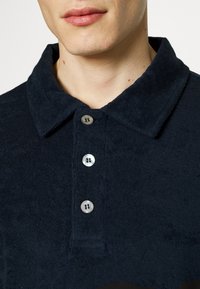 Närbild av en person som bär en mörk marinblå t-shirt i frotté med tre knappar och en vikkrage.