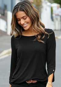 Schwarzes Langarmshirt mit rundem Ausschnitt und Knopfdetails an den Ärmeln, kombiniert mit einem geflochtenen braunen Gürtel, in einer lässigen Umgebung im Freien.