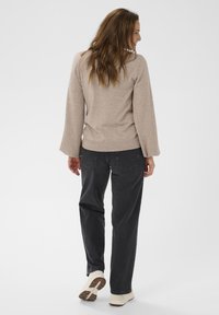 Pull marron clair avec manches longues évasées, associé à un jean gris foncé et des chaussures texturées de couleur claire. Les cheveux sont lâchés et ondulés.