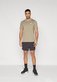 Tricou verde măsliniu, relaxat, cu logo alb, purtat cu șorturi negre. Modelul poartă pantofi sport negri și șosete până la glezne. Design simplu, sportiv.