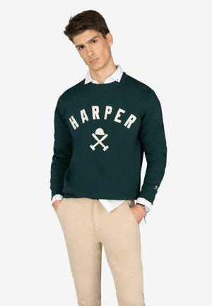 Sudadera verde con el logo "HARPER" y bates de béisbol cruzados en color crema; combinada con una camisa blanca con cuello y pantalones beige.