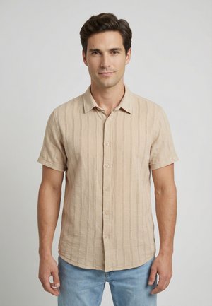 Homme aux cheveux courts et foncés portant une chemise à manches courtes beige à boutons et un jean bleu clair, debout devant un fond clair uni.