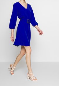 Blå klänning med V-ringning och puffärmar, åtdragna i midjan med en fyrkantig spänne. Buren med blommiga rosa klacksandaler.