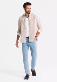 Camisa de botón color beige claro sobre una camiseta blanca, combinada con jeans azul claro y zapatos de barco oscuros, creando un atuendo casual.
