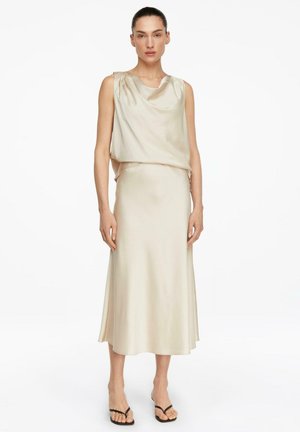 Kobieta ubrana w kremowy satynowy top bez rękawów i dopasowaną midi spódnicę, w czarnych sandałach na obcasie z odkrytymi palcami, stojąca na białym tle.