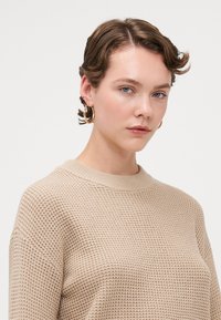 Beige strikkegenser med mønster, ribbestrikket rundhals og løs passform. Modellen har på seg gulløredobber, myk sminke og kort hår.