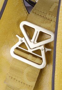 Szczegół żółtej skórzanej torby, z metaliczno-złotym trójkątnym klamrą oraz teksturowanym paskiem z subtelnym wzorem.