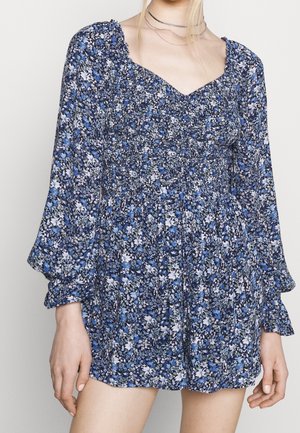 Kobieta w niebiesko-białej sukience z długim rękawem, ozdobionej kwiatowym wzorem, z marszczoną talią i dekoltem w kształcie serca, uzupełniona cienkimi srebrnymi naszyjnikami.