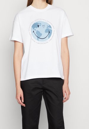 Osoba ubrana w biały T-shirt z niebieskim nadrukiem Ziemi przedstawiającym uśmiechnięte usta oraz sowę powyżej, w zestawieniu z czarnymi spodniami.