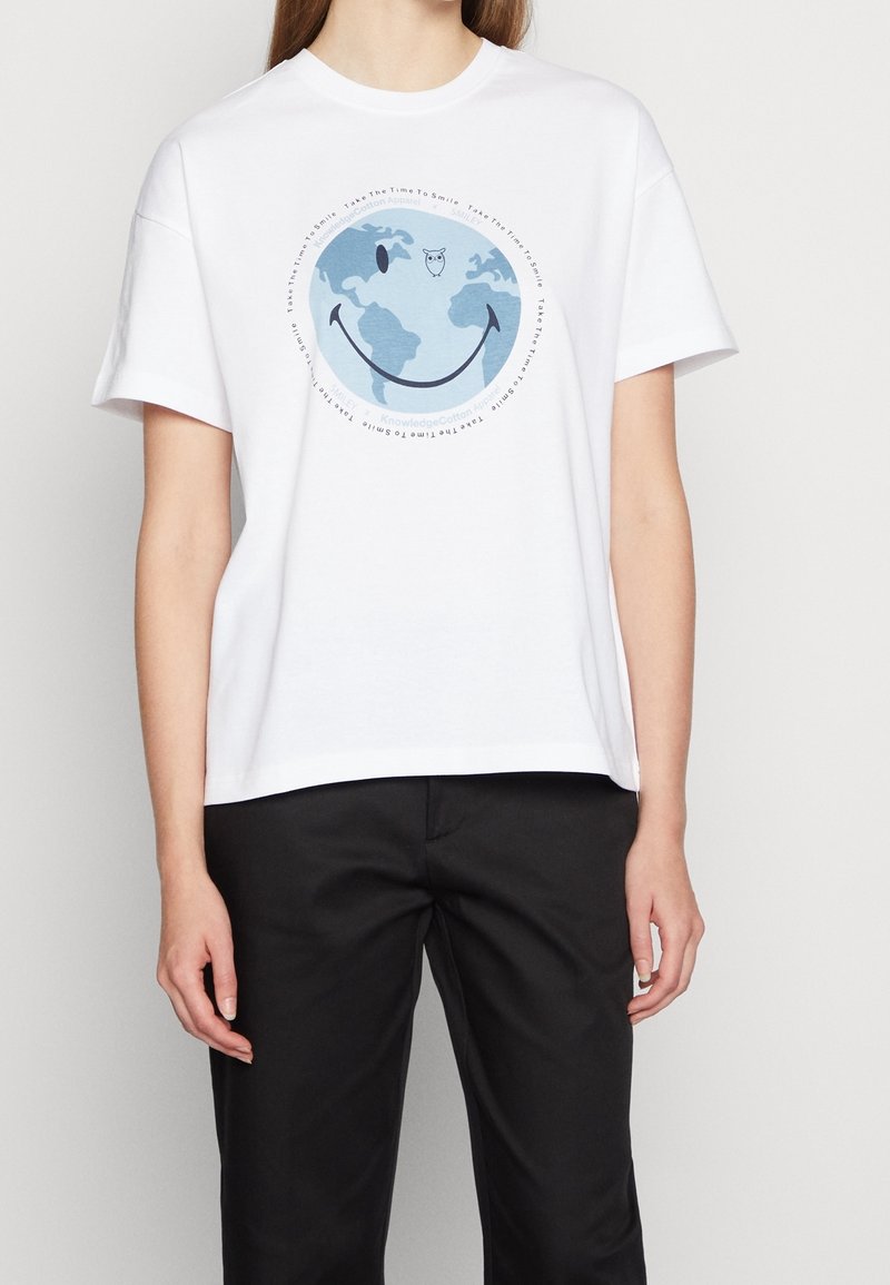 Personne portant un T-shirt blanc avec un graphique bleu de la Terre, affichant une bouche souriante et un hibou au-dessus, associé à un pantalon noir.