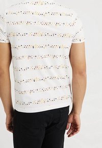 Valkoinen lyhythihainen t-paita, jossa on geometrinen kuvio, jossa on kolmioita sinisessä, oranssissa ja keltaisessa. Pehmeää kangasta, istuva malli.