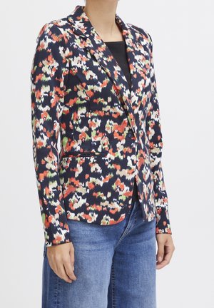 Femme portant un blazer ajusté à un seul bouton avec un imprimé floral abstrait multicolore et un jean bleu, debout devant un fond uni.