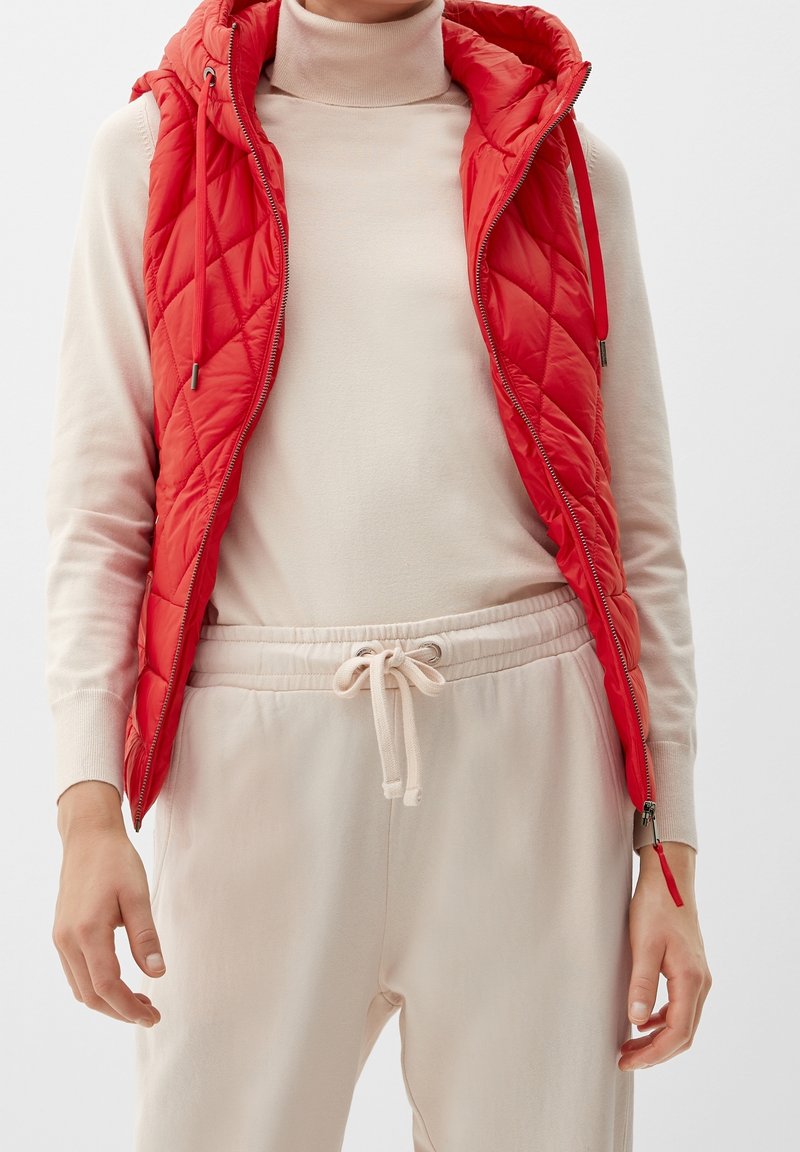 Gilet matelassé rouge avec capuche, doté d'une fermeture éclair et de cordons de serrage, porté sur un col roulé beige clair, associé à un pantalon de survêtement beige assorti.