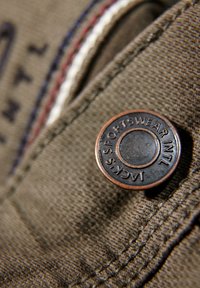 Bouton en métal sur tissu marron, avec le texte « JACK'S SPORTSWEAR INTL » en relief. Couture visible avec des accents rouges et blancs.