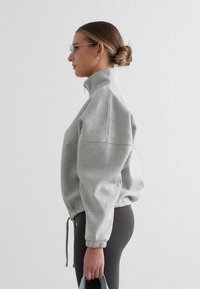 Lys grå zip-up genser laget av mykt stoff, med høy krage og elastiske mansjetter, kombinert med mørke leggings og holder en veske.