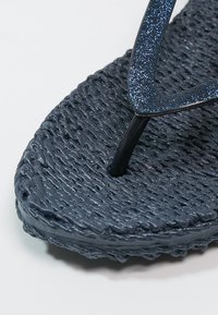 Infradito blu scuro con texture, cinturino a V scintillante e punta arrotondata su sfondo bianco.