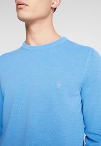 Pull bleu clair à col rond en tissu tricoté texturé. Présente un petit logo brodé sur la poitrine. Design simple et classique.