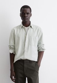 Striped button-up shirt in soft green and white. Includes a chest pocket, rolled sleeves, and a collared neckline, paired with dark trousers.