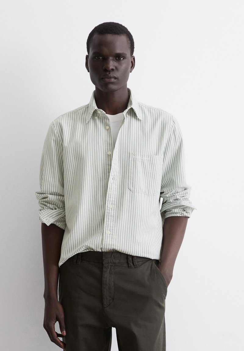 Striped button-up shirt in soft green and white. Includes a chest pocket, rolled sleeves, and a collared neckline, paired with dark trousers.
