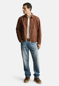 Bruine ritsjack, witte shirt eronder, lichtblauwe spijkerbroek, bruine schoenen met dikke zool. Model staat tegen een witte achtergrond.