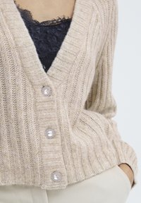 Cardigan beige à côtes fabriqué en maille douce, doté d'un décolleté en V profond, de trois boutons en cristal décoratifs et de poignets texturés.