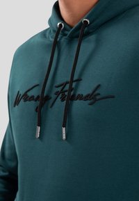 Felpa color teal realizzata in tessuto morbido con cappuccio e lacci neri, caratterizzata da un logo ricamato "Wrong Friends" sul petto.