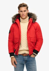 Chaqueta acolchada roja con capucha de pelo sintético desmontable, que cuenta con dos bolsillos frontales y puños negros acanalados, llevada sobre un suéter de punto trenzado color crema.