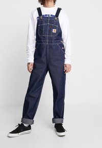 Tummansiniset farkkutypeille säädettävät olkaimet, etutasku, sivutaskut, kontrastiset tikkauskohdat, päällä valkoinen pitkähihainen paita.