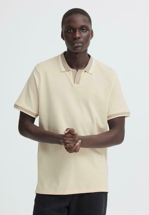 Giovane con la pelle scura che indossa una polo beige a maniche corte e pantaloni neri, in piedi con le mani intrecciate davanti.