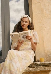 Femme en robe fleurie et bandeau en tricot lit un livre intitulé « Crudités » assise sur un rebord en pierre près d'une fenêtre avec une tasse jaune à proximité.