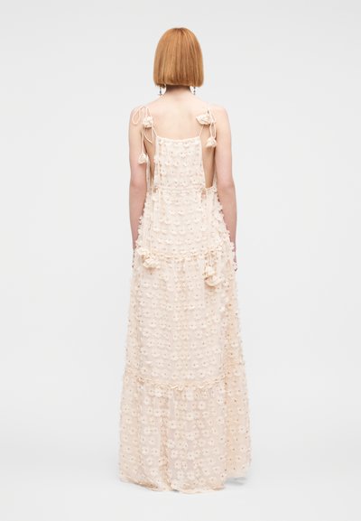 Robe longue beige pâle, en tissu léger, ornée de motifs floraux, bretelles spaghetti avec nœuds, et silhouette fluide.