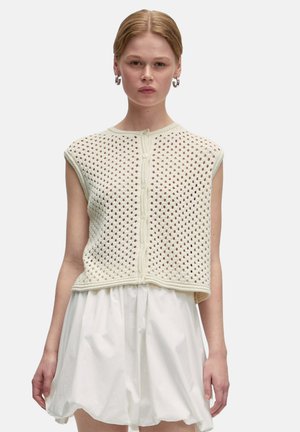 Gehaakte mouwloze top in crème met cirkelvormige patronen, voorzien van een ronde halslijn en een knoopsluiting aan de voorkant, gedragen met een witte rok.