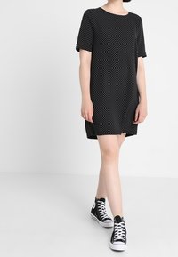 Schwarzes Kleid mit kurzen Ärmeln und weißen Punkten, aus leichtem Stoff, kombiniert mit schwarzen High-Top-Sneakern.