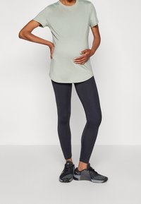 Camiseta de maternidad en verde claro, de manga corta, ajuste holgado; combinada con leggings negros y zapatillas deportivas negras con acentos texturizados.