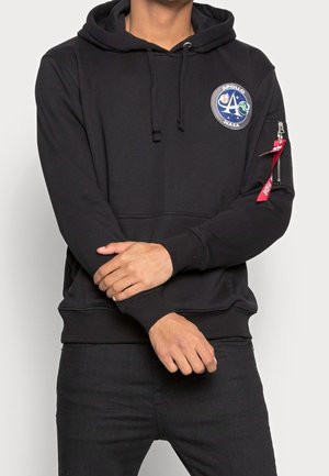 Osoba ubrana w czarną bluzę z kapturem NASA Apollo z naszywką na klatce piersiowej oraz czerwonym tagiem na kieszeni na zamek na lewym rękawie.