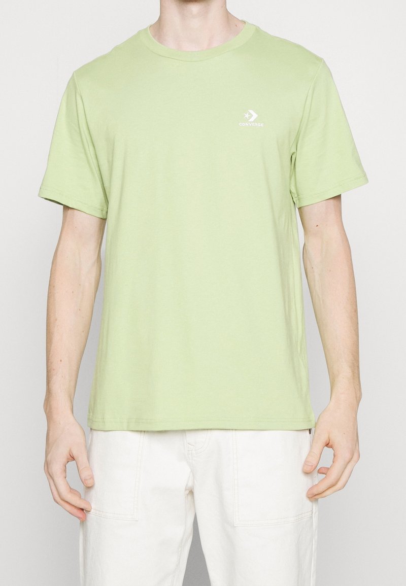 Homme portant un t-shirt Converse vert clair à manches courtes et un pantalon blanc, debout devant un fond clair uni.