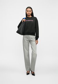 Černá mikina s růžovým textem, světle šedé džíny a černé lodičky. Modelka drží černou tašku a stojí na jednobarevném pozadí.