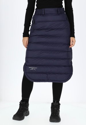 Navy puffer skirt with horizontal quilted pattern, mid-calf length, featuring side zippers and a logo at the hem. Worn over black leggings and boots.