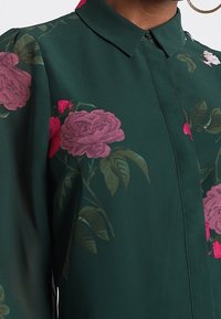 Blouse vert foncé avec un motif floral rose, dotée d'un col pointu et de boutons dissimulés à l'avant, portée avec une grande boucle d'oreille en or.