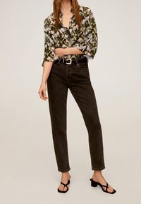 Donna che indossa una blusa floreale verde e rosa, jeans neri a vita alta con cintura e sandali con tacco neri, in piedi con un braccio incrociato.