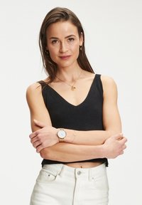 Zwarte geribbelde fitted tanktop, gouden horloge met een witte wijzerplaat en gelaagde gouden sieraden. Model heeft lang bruin haar en gekruiste armen.