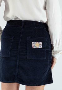 Marineblauer Cordrock mit taillierter Passform, ausgestattet mit aufgesetzten Taschen und einem dekorativen Etikett mit Blumenmuster auf der Rückentasche.
